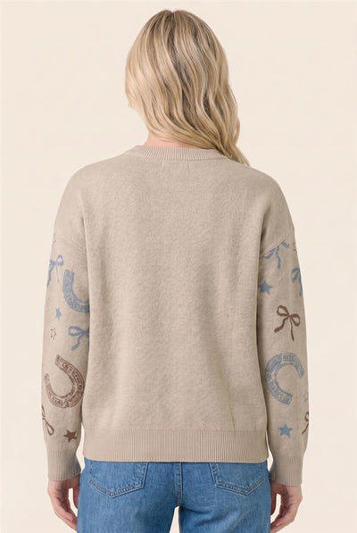 A woman viewed from behind wearing a light beige knit sweater with western-themed embroidery on both sleeves, paired with blue jeans, against a solid light beige background.