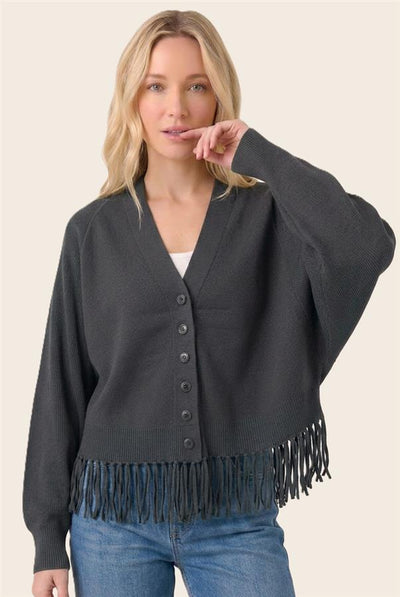 A woman wearing a dark charcoal gray knit cardigan with fringe trim at the hem and button-front closure, paired with blue jeans, against a solid light beige background.