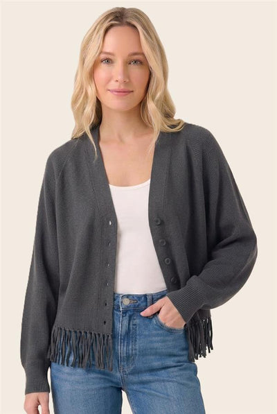 A woman wearing a dark gray knit cardigan with fringe trim over a white tank top and blue jeans, photographed against a plain light beige studio background.