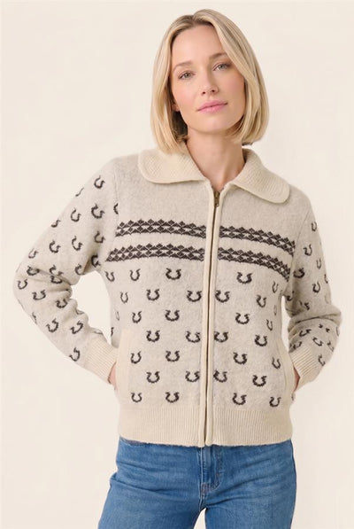 A woman wearing a cream-colored zip-up cardigan with black horseshoe patterns and dark horizontal stripes, paired with blue jeans, against a plain beige background.