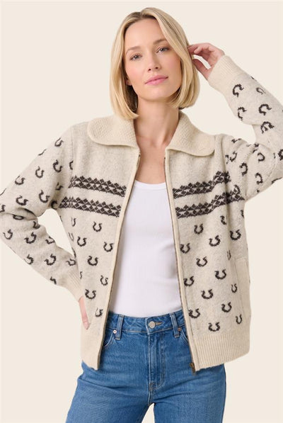 A woman wearing a cream-colored knit zip-up cardigan with black horseshoe and geometric pattern details, styled over a white tank top and blue jeans against a light beige background.