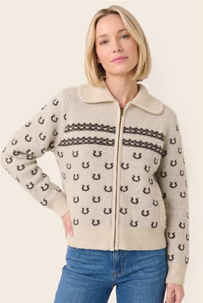 A woman wearing a beige knit zip-up cardigan with a dark brown horseshoe pattern and horizontal decorative bands, paired with blue jeans, against a solid light beige background.