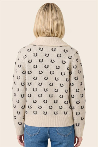 A woman viewed from the back wearing a cream knit sweater with a repeating black horseshoe pattern and blue jeans, against a plain light beige studio background.