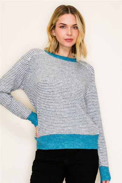 A woman wearing a long-sleeve knit sweater with fine horizontal gray and white stripes, featuring solid turquoise blue ribbed trim at the crew neck, cuffs, and hem, paired with black pants against a plain off-white background.
