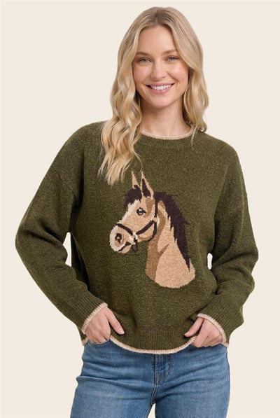 A woman wearing an olive green knit sweater with a large horse head graphic on the front, paired with blue jeans, against a solid light beige background.