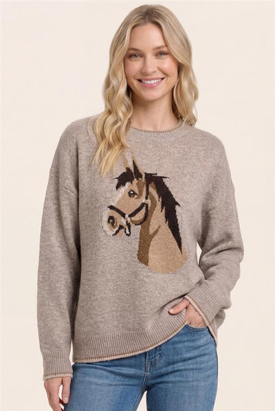 A woman wearing a taupe knit sweater with a large horse head graphic on the front, paired with blue jeans, against a solid light beige background.