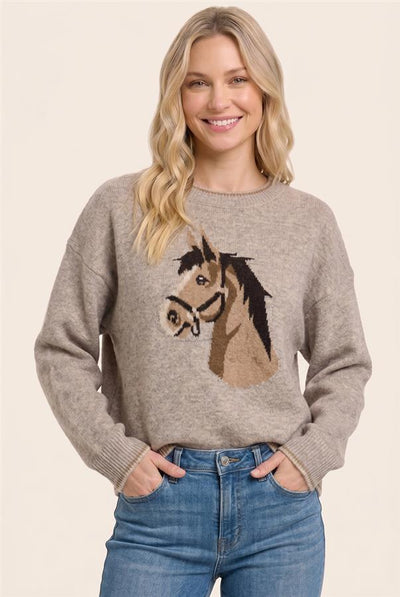 A woman wearing a light taupe knit sweater with a knitted horse head graphic on the front, paired with blue jeans, against a solid light beige background.