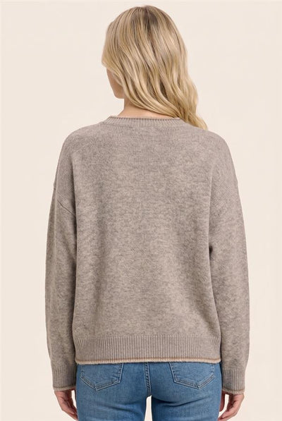 A woman viewed from the back wearing a light taupe knit sweater and blue jeans against a solid cream background.