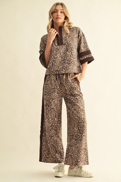 A woman modeling a matching two-piece lounge set with a brown and beige leopard print pattern, featuring a short-sleeve top with dark brown trim and wide-leg pants with side panels, paired with white sneakers against a plain beige background.