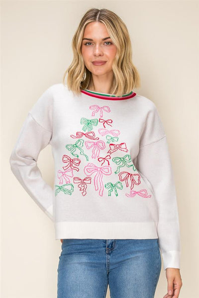 A woman wearing a white crewneck sweatshirt decorated with a colorful embroidered Christmas tree made of pink and green bows, paired with blue jeans, against a solid beige background.