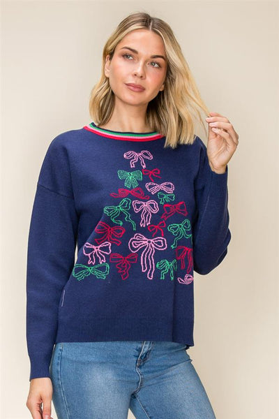 A woman wearing a navy blue crewneck sweater with a colorful embroidered Christmas tree made of bows in pink, green, and purple, paired with blue jeans, against a plain beige background.