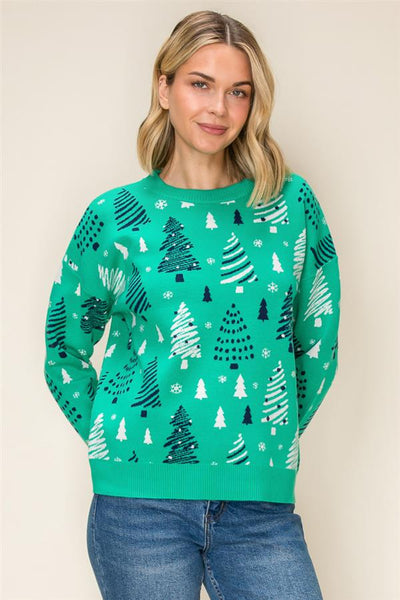 A woman wearing a festive green crewneck sweater with a white and dark green Christmas tree pattern, paired with blue jeans, set against a plain beige background.