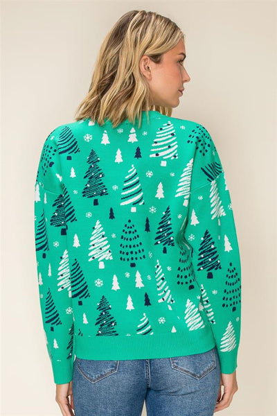 A woman shown from the back wearing a bright green sweater with an all-over pattern of stylized Christmas trees in dark green and white, paired with blue jeans against a plain beige background.