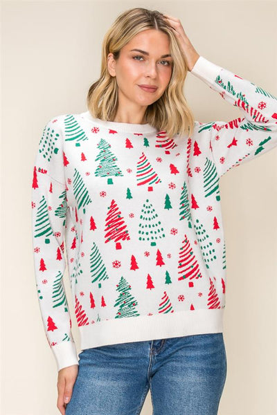 A woman wearing a white crewneck sweater with a festive Christmas tree pattern in red and green, paired with blue jeans, against a solid beige background.