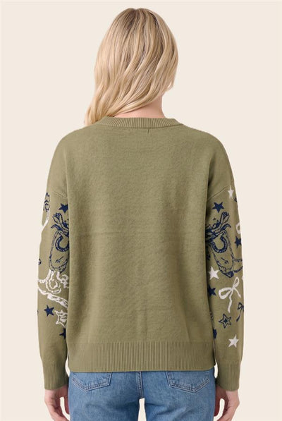 A woman viewed from the back wearing an olive green knit sweater with navy and white celestial embroidery on the sleeves, paired with blue jeans against a light beige background.