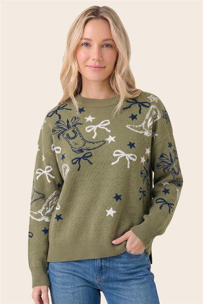 A woman wearing an olive green knit sweater with a whimsical pattern of white and navy blue stars, bows, and sleighs, paired with blue jeans against a solid light beige background.