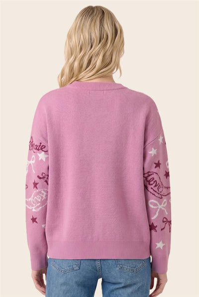 A woman viewed from behind wearing a pink knit sweater with white and dark pink embroidered script and star motifs on the sleeves, paired with blue jeans against a light beige background.