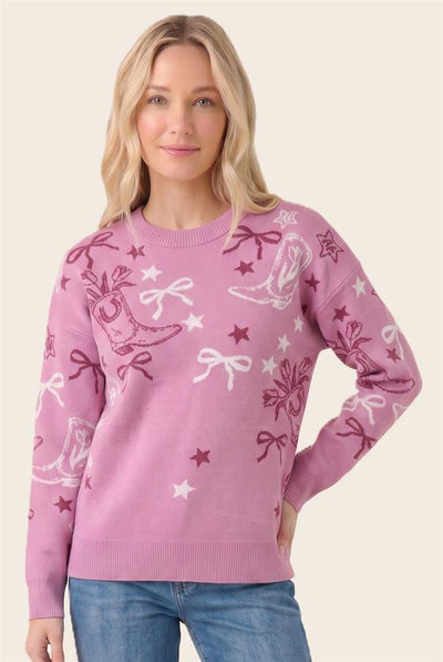 A woman wearing a pink crewneck sweater with a festive knit pattern of white and dark pink boots, bows, stars, and reindeer, paired with blue jeans against a solid light beige background.