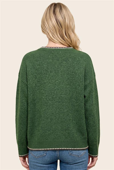 A woman viewed from the back wearing a dark green knit sweater with decorative white and black trim along the neckline and hem, paired with blue jeans against a light beige background.