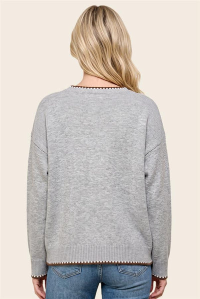 A woman viewed from the back wearing a light gray knit sweater with decorative brown pom-pom trim along the neckline, cuffs, and hem, paired with blue jeans against a solid beige background.