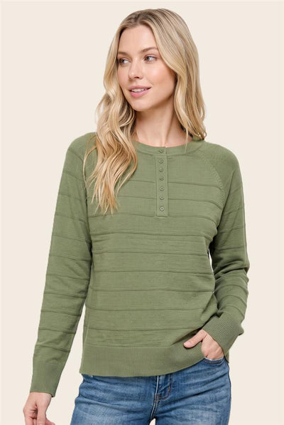 A woman wearing an olive green ribbed long-sleeve henley sweater with button details on the chest, paired with blue jeans, against a solid beige background.