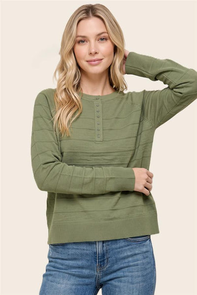 A woman wearing an olive green ribbed long-sleeve henley sweater with button details on the chest, paired with blue jeans, against a solid beige background