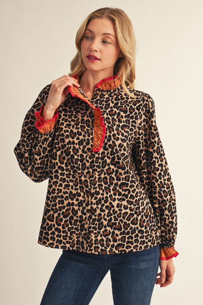 A woman modeling a leopard-print long-sleeve blouse with a ruffled collar and cuffs in orange-red, paired with blue jeans against a plain beige studio background.