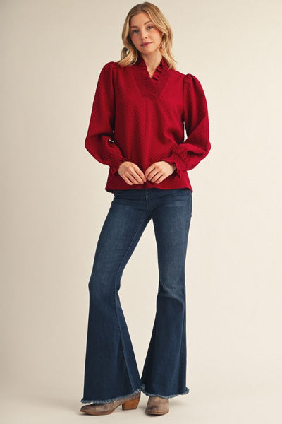 A woman modeling a deep red long-sleeve blouse with puffed shoulders and a ruffled collar, paired with dark blue flare jeans and brown heeled boots, against a plain beige studio background.