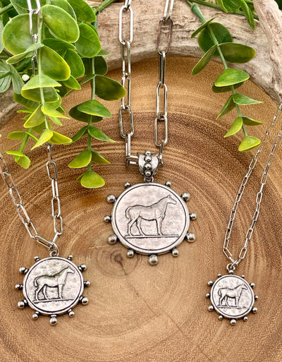 Three silver necklaces with horse-themed pendants on a wooden surface with green leaves.
