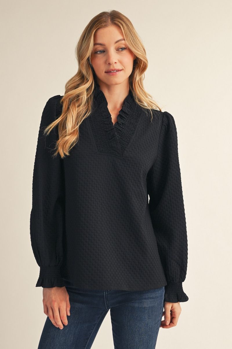 A woman modeling a black long-sleeve blouse with a textured waffle pattern, ruffled V-neckline, and puffed sleeves, paired with blue jeans against a plain beige background.
