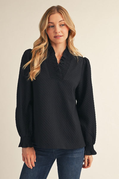 A woman modeling a black long-sleeve blouse with a textured waffle pattern, ruffled V-neckline, and puffed sleeves, paired with blue jeans against a plain beige background.