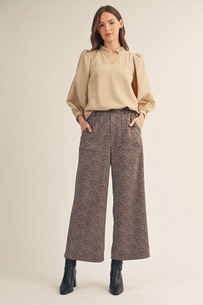 A woman modeling a beige long-sleeve blouse and brown patterned wide-leg trousers with black ankle boots, standing against a plain light beige studio background.