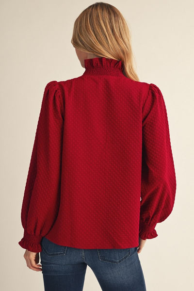 A woman viewed from the back wearing a textured red long-sleeve blouse with a ruffled mock neck and puffed sleeves, paired with blue jeans against a plain beige background.