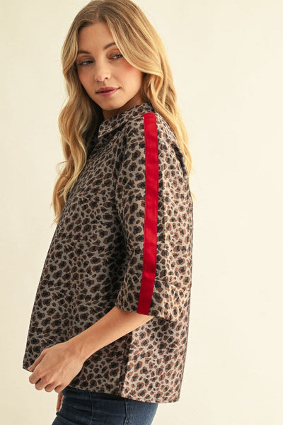 A woman wearing a leopard print short-sleeve button-up shirt with a bold red vertical stripe down the sleeve, paired with dark blue jeans, set against a plain cream background.