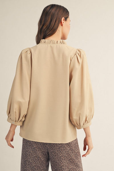 A woman shown from the back wearing a beige blouse with a ruffled high neck and voluminous elbow-length puff sleeves, paired with leopard-print pants against a plain light beige studio background.