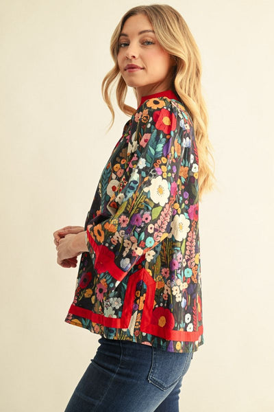 A woman wearing a colorful floral print blouse with red trim, paired with dark blue jeans, photographed against a plain light beige background.