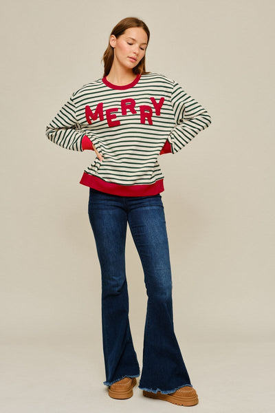 A woman modeling a holiday-themed long-sleeve sweater with green and white horizontal stripes, red trim, and 'MERRY' written across the chest in red, paired with dark blue flared jeans and brown boots, against a solid beige studio background.
