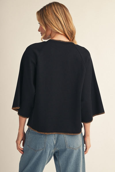 A woman shown from the back wearing a black cropped top with wide three-quarter sleeves and tan contrast stitching along the edges, paired with light blue jeans against a plain beige studio background.