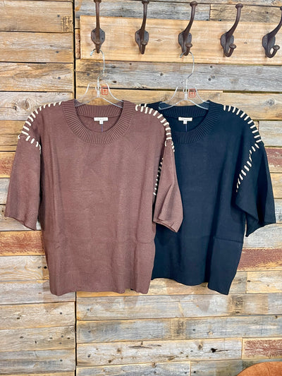 Two short-sleeve knit sweaters hang on hangers against a rustic wooden plank wall. The sweater on the left is a brown color with white contrast stitching along the shoulders, and the sweater on the right is black with matching white contrast stitching along the shoulders.