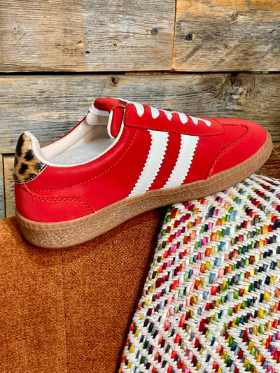 A red leather sneaker with three white stripes, a brown jute espadrille sole, and a leopard-print heel tab, resting on a brown leather surface next to a colorful woven textile against a rustic wood plank background.
