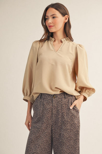 A woman wearing a beige long-sleeve blouse with a ruffled V-neck collar and puff sleeves, paired with brown leopard-print trousers, against a plain light beige studio background.