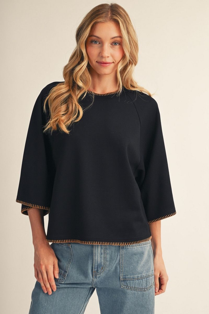 A woman wearing a black three-quarter sleeve top with gold trim on the collar, hem, and cuffs, paired with light blue jeans, against a plain beige background.