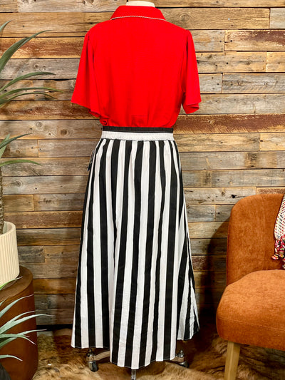 Mannequin wearing a red shirt and black and white striped skirt against a wooden wall.