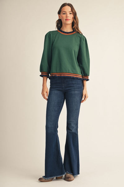 A woman modeling a dark green long-sleeve knit top with orange and navy striped trim at the crew neck, cuffs, and hem, paired with dark blue flare jeans and brown shoes, against a solid beige studio background.