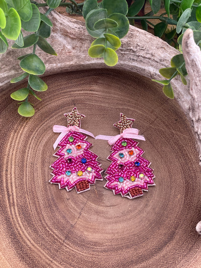 Pink Christmas tree earrings on a wooden surface with green leaves.