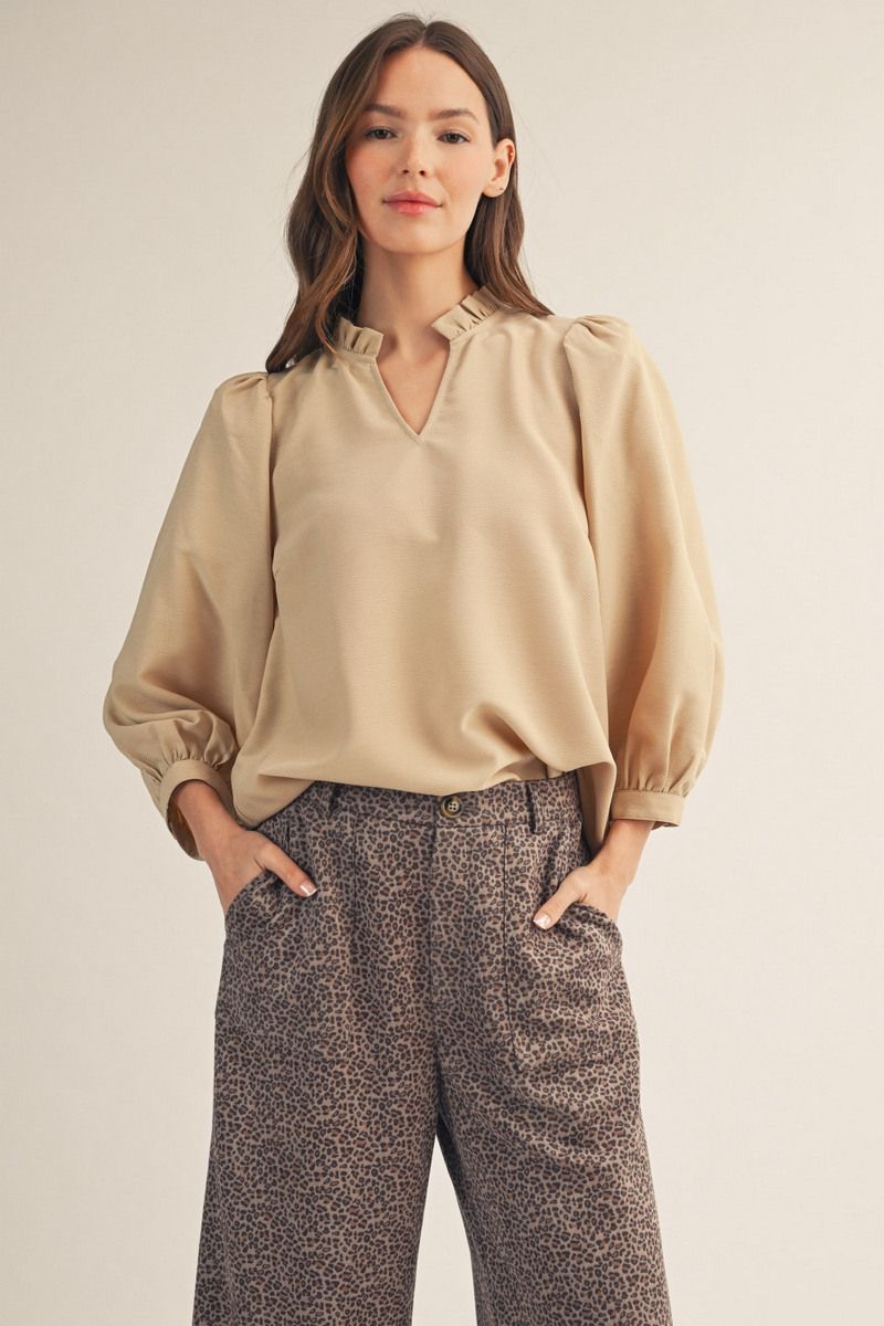 A woman modeling a beige long-sleeve blouse with a ruffled V-neck collar and gathered cuffs, paired with brown and beige patterned trousers, against a plain light beige studio background.