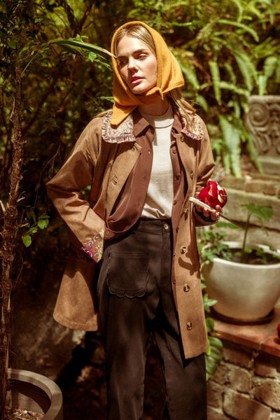 A woman stands outdoors in a garden setting wearing a brown double-breasted coat with ornate patterned collar, a mustard yellow headscarf, a white top, and dark brown wide-leg trousers, while holding a red apple.