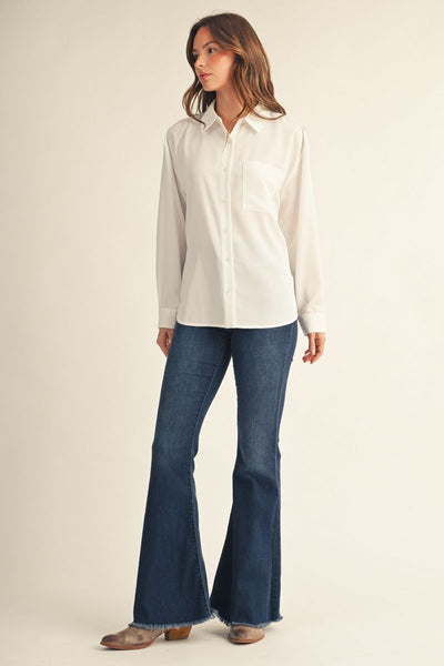 A woman modeling a white long-sleeve button-up shirt with a chest pocket, paired with dark blue flared jeans and brown boots, against a plain beige studio background.