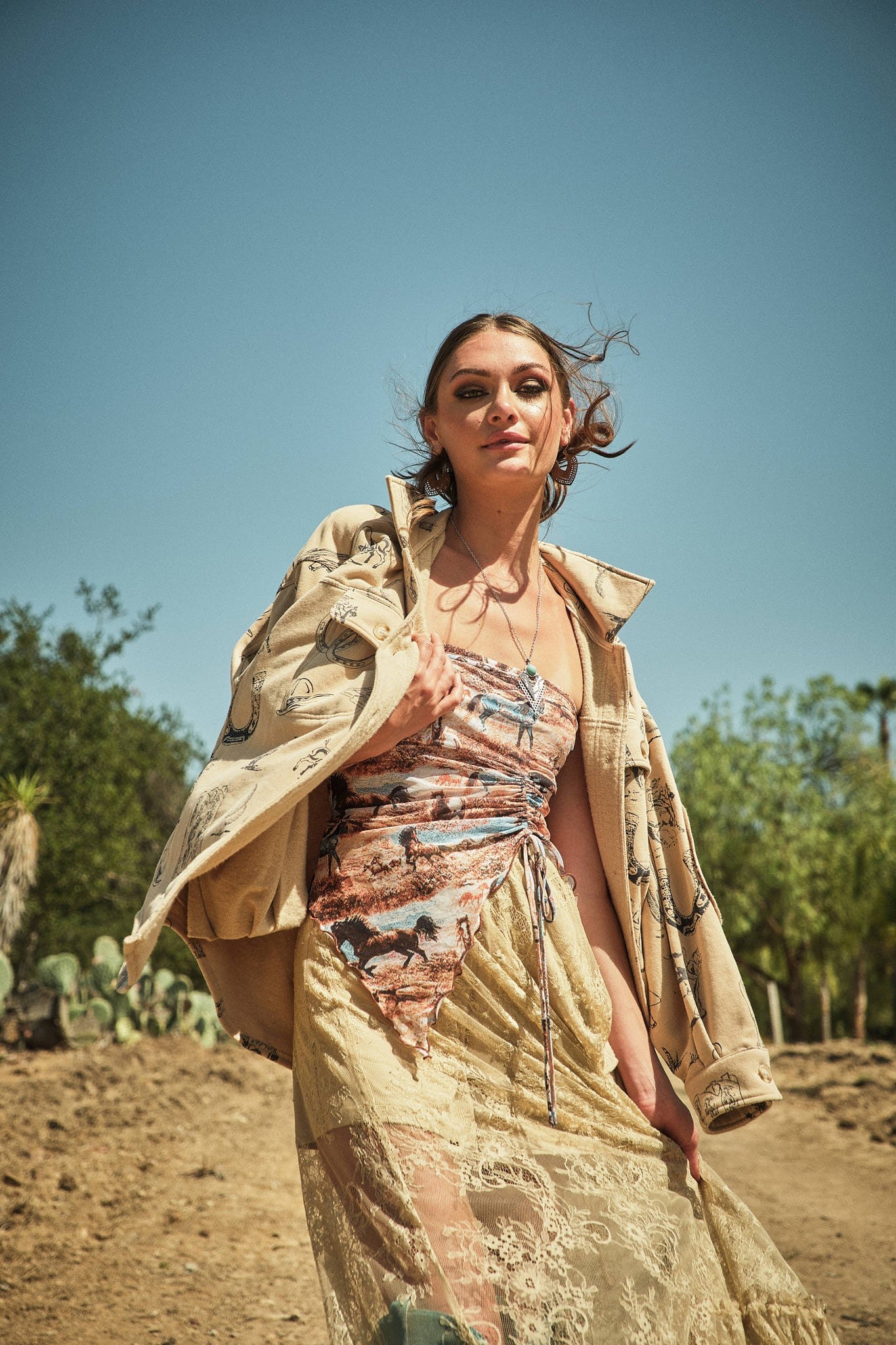 Woman in a patterned dress and beige jacket standing in a desert landscape with trees in the background.