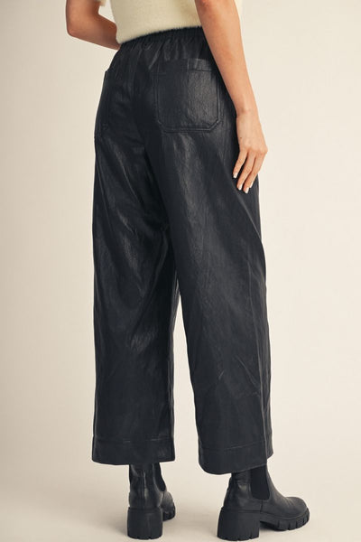 Black wide-leg faux leather pants with a back pocket, shown on a model from the back wearing black Chelsea boots against a light beige studio background.